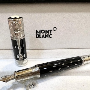 May include: A Montblanc fountain pen with a black barrel and white polka dots. The pen features a silver-colored nib and accents, and the cap is off to the side. The pen is in a white box with the Montblanc logo.
