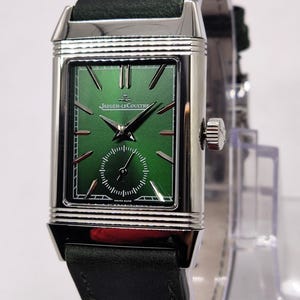 May include: A rectangular, silver wristwatch with a green face and a black leather strap. The watch features a small seconds dial and the Jaeger-LeCoultre logo. The watch strap is dark green.