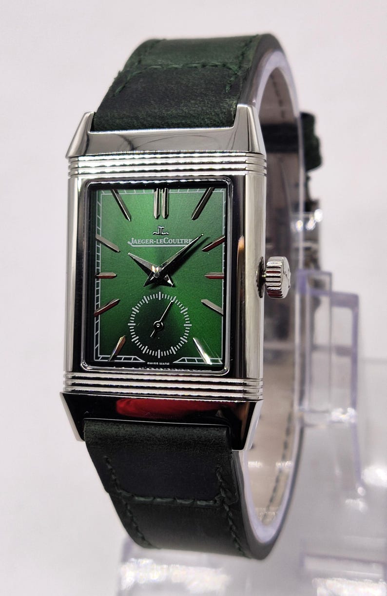 May include: A rectangular, silver wristwatch with a green face and a black leather strap. The watch features a small seconds dial and the Jaeger-LeCoultre logo. The watch strap is dark green.