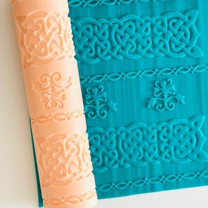 May include: A cylindrical peach-coloured rolling pin with an engraved pattern of Celtic knots and floral motifs, positioned next to a flat sheet of teal material that displays the same embossed designs in a repeating horizontal layout.