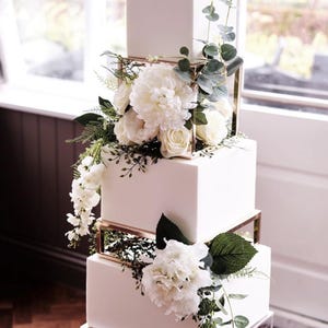 May include: A four-tier square white cake featuring metallic gold-toned geometric separators between the tiers. The cake is decorated with white floral arrangements including peonies, hydrangeas, and eucalyptus greenery. The base is a clear stand.
