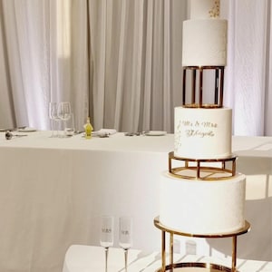 May include: A tall, multi-tiered white wedding cake with gold detailing and a gold stand. The cake features a textured pattern and gold lettering that reads "Mr. & Mrs. W. Bugego". Two champagne flutes with "MRS" and "MR" are in the foreground.