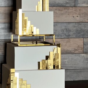 May include: A four-tiered square cake with smooth white icing, featuring reflective gold rectangular accents arranged in ascending steps on each tier. A gold metal frame separates the second and third tiers of this multi-level cake.