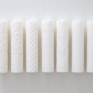 May include: Seven white cylindrical vases with embossed floral and geometric patterns. Each vase has a unique design, including swirls, leaves, and circles. The vases are arranged in a row against a plain white background.