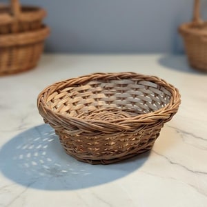 Pode incluir: Uma pequena cesta ovalada tecida com vime castanho claro. A cesta tem uma superfície texturizada e uma borda arredondada. Outras cestas são visíveis no fundo, sugerindo um conjunto ou coleção.