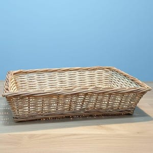 May include: A rectangular wicker basket in a light brown colour. The empty basket sits on a light wood surface against a blue background. The basket's woven design is clearly visible.