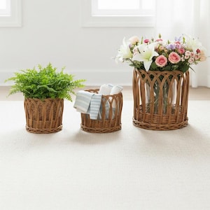 Puede incluir: Tres cestas de mimbre tejidas de diferentes tamaños. Una contiene una planta verde, otra toallas dobladas y la más grande un ramo de flores. Las cestas son marrones y están sobre una alfombra clara.