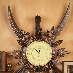 Puede incluir: Un reloj de pared decorativo de madera con una esfera de color crema y la palabra "Ajanta" impresa. El reloj está rodeado por un diseño de sol con detalles tallados y cristales incrustados. Las manecillas del reloj señalan las 12:00.