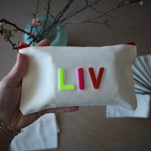 Könnte beinhalten: Eine cremefarbene Canvas-Tasche mit dem Namen "LIV" in neongrünen, rosa- und orangefarbenen Buchstaben. Die Tasche hat einen roten Reißverschluss und wird in einer Hand gehalten. Ein goldenes Armband ist am Handgelenk zu sehen.