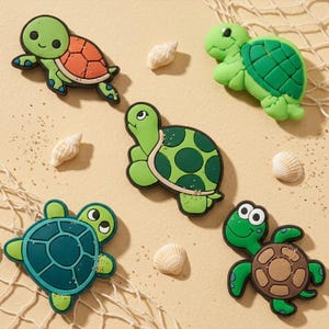 May include: Five colorful turtle-shaped magnets in various shades of green, brown, and teal. The turtles have detailed shells and friendly faces. Small seashells are scattered around the magnets on a sandy surface, with a fishing net in the background.