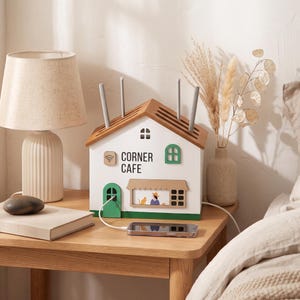Puede incluir: Una cubierta para router en forma de casa blanca con techo de madera y cuatro ranuras verticales. El frente presenta una puerta verde, una ventana verde, un icono de Wi-Fi y el texto CORNER CAFE sobre un gráfico de una persona y un gato.
