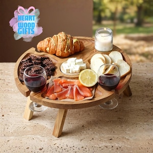 May include: A wooden picnic table with a round serving tray. The tray holds food items like a croissant, cheese, crackers, salmon, and bread. Two wine glasses are on the tray, and a candle is in a jar. The table has three legs.