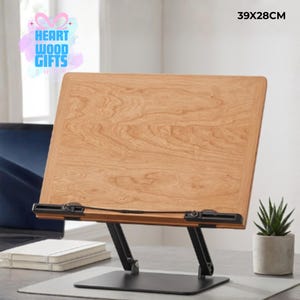 May include: A wooden book stand, 39 x 28 cm, with a light brown finish. It features a black metal base and adjustable angle settings. The Heart Wood Gifts Shop logo is visible. A small succulent plant in a concrete pot is on the right.