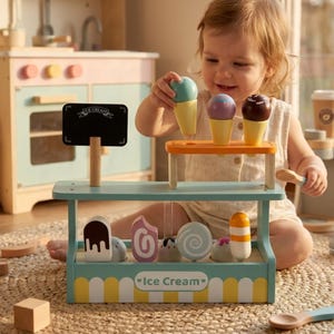 May include: A wooden ice cream stand with a chalkboard sign and various ice cream cones and treats. The stand is painted in pastel colors with a yellow and white scalloped trim. The sign reads "Ice Cream".
