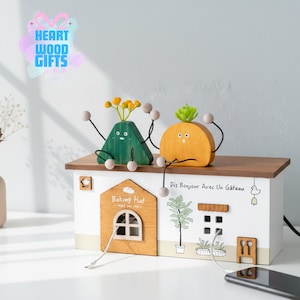 May include: A whimsical wooden charging station shaped like a house, featuring a brown roof and white walls. Two cartoon-like figures sit atop the roof. The house has a "Baking Hut" sign and the text "Dis Bonjour Avec Un Gâteau". A phone rests nearby.