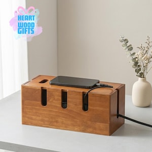 May include: A rectangular wooden cable organizer with a phone on top. The organizer has slots for cords and a hole for the charging cable. The wood has a warm, natural finish. The product is from Heart Wood Gifts Shop.