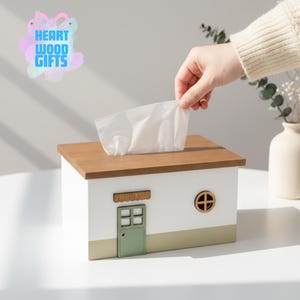 May include: A white tissue box shaped like a house with a brown roof, a green door, and a round window. The tissue box has the word "WELCOME" above the door. A hand is pulling a tissue from the box.
