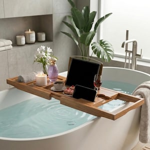 Puede incluir: Una bandeja de baño de madera sostiene una tableta, un teléfono, café, postre y una copa de vino. Una vela encendida, flores y un frasco de perfume crean un ambiente de spa relajante. La bandeja está sobre una bañera blanca llena de agua.