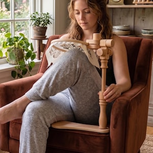 May include: A light-colored wooden embroidery hoop stand with an adjustable arm and a circular frame holding a piece of white fabric with a printed design. The stand features a flat base that rests on a seat and a vertical post with rotating joints.