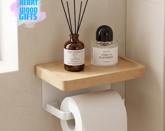 May include: A wooden shelf with a white toilet paper holder. The shelf holds a brown fragrance oil bottle with reeds and a clear bottle with a black cap. The text "FRAGRANCE OIL" and "STORY OF FLAVORIST" are visible on the bottles. The logo "HEART WOOD GIFTS" is in the upper left corner.