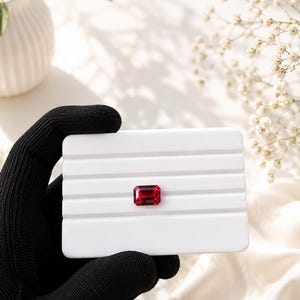 May include: A vibrant, rectangular, emerald-cut red gemstone centred on a white, rectangular display stand with horizontal grooves. The gemstone exhibits deep crimson tones and clear facets, contrasting against the smooth, matte white surface.