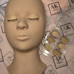 May include: A mannequin head with closed eyelids and applied false eyelashes. Several sets of false eyelashes are visible in a clear plastic container. The background features printed tissue paper with the text "AK BEAUTY ANNIE KEY".