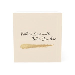 Puede incluir: Una caja de color crema con el texto "Fall in Love with Who You Are" en escritura negra. Debajo del texto hay una pincelada dorada.
