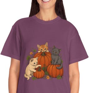 Peut inclure: Un t-shirt à manches courtes de couleur prune avec une impression graphique représentant trois chatons interagissant avec des citrouilles orange. Un chaton tigré orange est assis sur une citrouille, un chaton crème touche une citrouille.