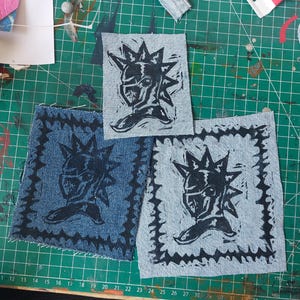 May include: Three rectangular denim patches featuring a black ink-printed graphic of a stylised knight helmet with a spiked crest. Two of the patches include a decorative barbed wire-style border frame around the central knight helmet illustration.