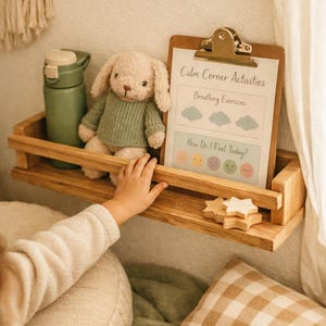 Könnte beinhalten: Ein Holzregal mit einer grünen Wasserflasche, einem Stoffhasen mit grünem Pullover und einem Klemmbrett mit dem Text "Calm Corner Activities". Das Regal enthält auch Holzstern-Dekorationen. Eine Kinderhand greift nach dem Regal.