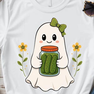 Op de afbeelding: Een wit t-shirt met een cartoon-geest met een groene strik op het hoofd, die een glazen pot met groene augurken vasthoudt. De geest wordt geflankeerd door twee gele bloemen met groene stengels en bladeren, in een eenvoudige grafische stijl.