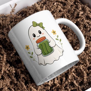May include: A white ceramic mug featuring a cartoon illustration of a ghost wearing a green bow and holding a glass jar filled with green pickles. The design includes small yellow flowers on either side of the ghost on a white background.