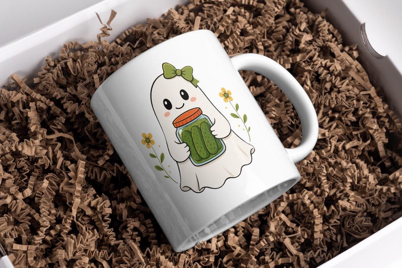 May include: A white ceramic mug featuring a cartoon illustration of a ghost wearing a green bow and holding a glass jar filled with green pickles. The design includes small yellow flowers on either side of the ghost on a white background.