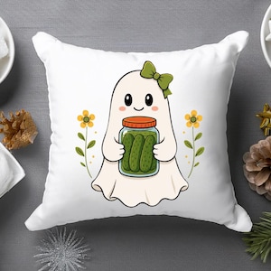 May include: A white square throw pillow featuring a central graphic of a cartoon ghost with a green bow, holding a glass jar filled with green pickles. The ghost is flanked by two small yellow flowers on thin green stems, all set against a white background.