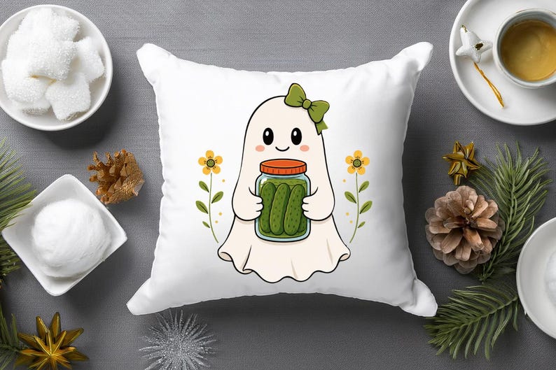 May include: A white square throw pillow featuring a central graphic of a cartoon ghost with a green bow, holding a glass jar filled with green pickles. The ghost is flanked by two small yellow flowers on thin green stems, all set against a white background.