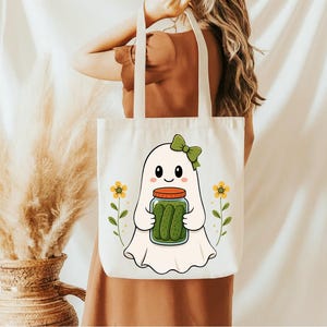 May include: A white canvas tote bag featuring a graphic illustration of a white ghost with a green bow, holding a glass jar filled with green pickles. The design is flanked by two small yellow flowers on thin green stems, centered on the bag.