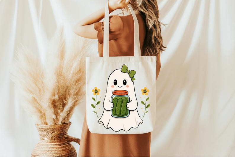 May include: A white canvas tote bag featuring a graphic illustration of a white ghost with a green bow, holding a glass jar filled with green pickles. The design is flanked by two small yellow flowers on thin green stems, centered on the bag.