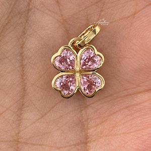 Puede incluir: Un colgante de oro con forma de trébol de cuatro hojas, con cuatro piedras preciosas rosas en forma de corazón en el centro de cada hoja. El colgante incluye una pequeña anilla de oro en la parte superior para sujetarlo a una cadena.