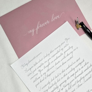 Może przedstawiać: Zakurzona różowa koperta z białym kursywnym napisem my forever love oraz biały list z odręczną kaligrafią, w towarzystwie czarnego pióra wiecznego ze złotą stalówką spoczywającego na rogu koperty.