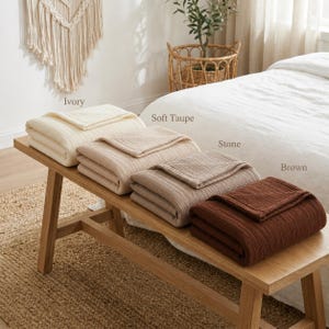 Pu&ograve; includere: Quattro coperte piegate, avorio, talpa chiaro, pietra e marrone, sono esposte su una panca di legno. Le coperte hanno una trama a coste. La panca &egrave; in legno chiaro e poggia su un tappeto tessuto.
