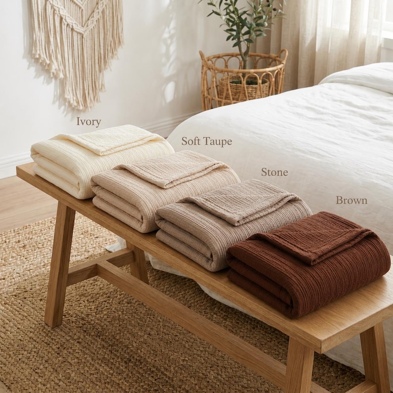 Pu&ograve; includere: Quattro coperte piegate, avorio, talpa chiaro, pietra e marrone, sono esposte su una panca di legno. Le coperte hanno una trama a coste. La panca &egrave; in legno chiaro e poggia su un tappeto tessuto.