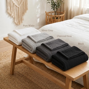 Pu&ograve; includere: Quattro coperte piegate in bianco puro, grigio argento, grigio antracite e nero sono esposte su una panca di legno. Le coperte hanno un design strutturato e a coste. I nomi dei colori sono stampati sopra ogni coperta.