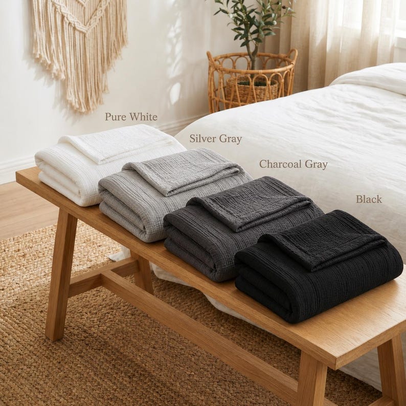 Pu&ograve; includere: Quattro coperte piegate in bianco puro, grigio argento, grigio antracite e nero sono esposte su una panca di legno. Le coperte hanno un design strutturato e a coste. I nomi dei colori sono stampati sopra ogni coperta.