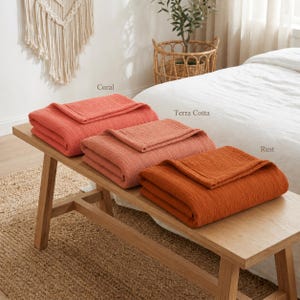 Pu&ograve; includere: Tre coperte piegate nei colori corallo, terracotta e ruggine sono esposte su una panca di legno. Le coperte hanno un aspetto strutturato. I nomi dei colori sono scritti sopra ogni coperta.