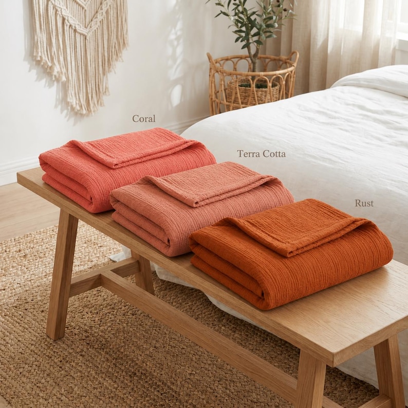 Pu&ograve; includere: Tre coperte piegate nei colori corallo, terracotta e ruggine sono esposte su una panca di legno. Le coperte hanno un aspetto strutturato. I nomi dei colori sono scritti sopra ogni coperta.