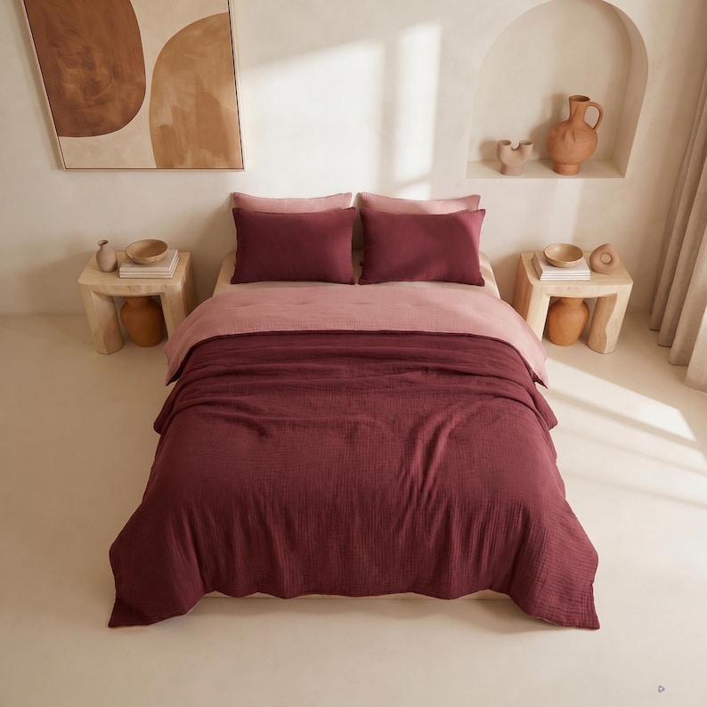 Pu&ograve; includere: Un letto con un piumino bordeaux e due federe abbinate. Il letto ha un lenzuolo rosa chiaro e due cuscini bordeaux. La stanza ha due piccoli comodini in legno con oggetti decorativi e un dipinto.