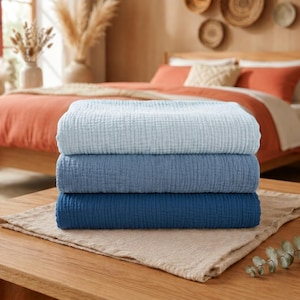 May include: A stack of three folded blankets featuring a textured waffle-weave pattern. The top blanket is light blue, the middle blanket is medium blue, and the bottom blanket is a deep navy blue, all resting on a beige fabric surface.