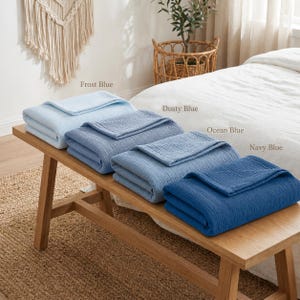 Pu&ograve; includere: Quattro coperte piegate in diverse tonalit&agrave; di blu, tra cui Frost Blue, Dusty Blue, Ocean Blue e Navy Blue, sono accuratamente impilate su una panca di legno. Le coperte hanno un aspetto strutturato e sono etichettate con i rispettivi colori.