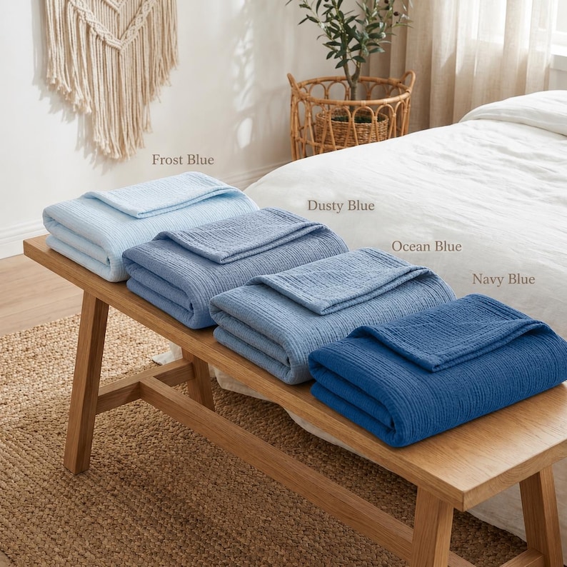 Pu&ograve; includere: Quattro coperte piegate in diverse tonalit&agrave; di blu, tra cui Frost Blue, Dusty Blue, Ocean Blue e Navy Blue, sono accuratamente impilate su una panca di legno. Le coperte hanno un aspetto strutturato e sono etichettate con i rispettivi colori.