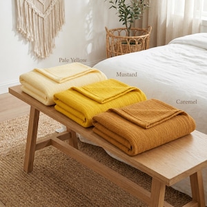 Pu&ograve; includere: Tre coperte piegate nei colori giallo pallido, senape e caramello sono esposte su una panca di legno. Le coperte hanno un design strutturato e a coste. La panca &egrave; posizionata su un tappeto tessuto, con un letto e oggetti decorativi sullo sfondo.